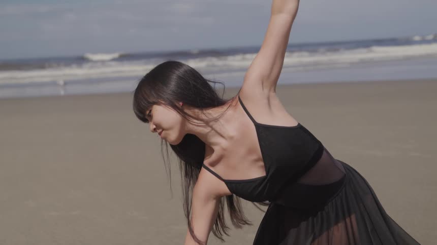 An asian dancer in a black dress is dancing gracefully on a beach