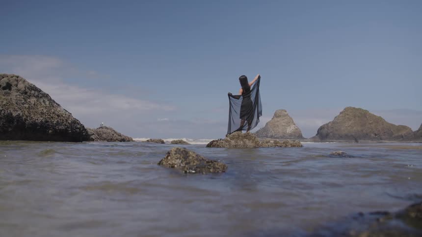 A woman dressed in black poses on a rock on a beach