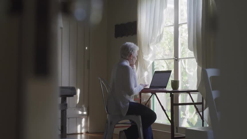 Woman with white hair sits at her desk on her laptop in her home