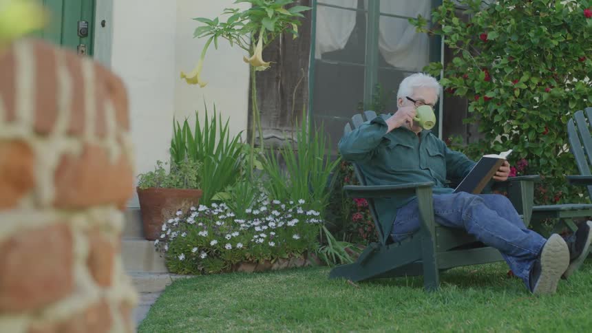 An elderly man sits outside reading a book and drinking out of a mug