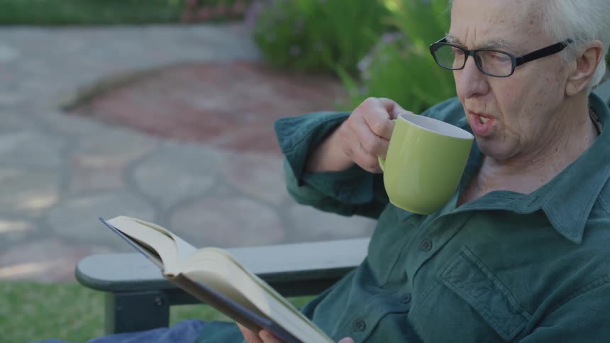 A man reading a book outside reaches for a mug on the armrest