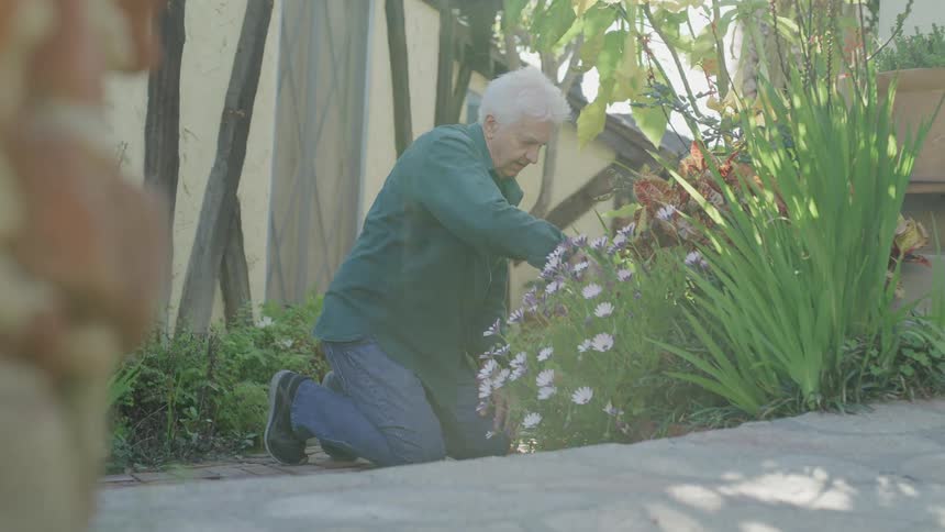 An old woman in a green shirt is on her knees working with flowers in her garden