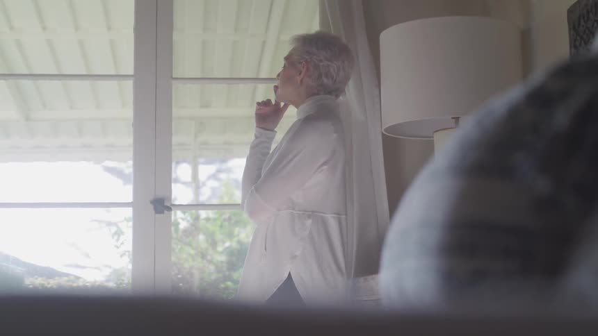 Old woman standing next to the bed looks out the window in the bedroom