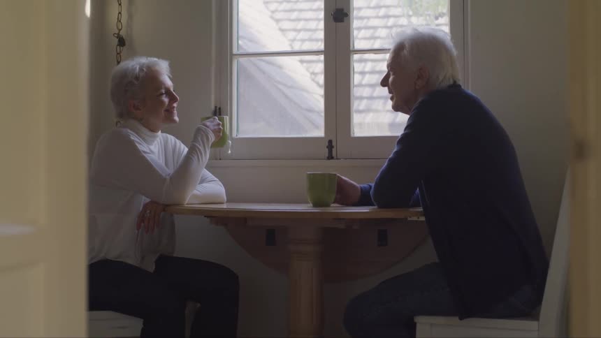An elder man and woman sit at a small table indoors as they converse and drink from mugs
