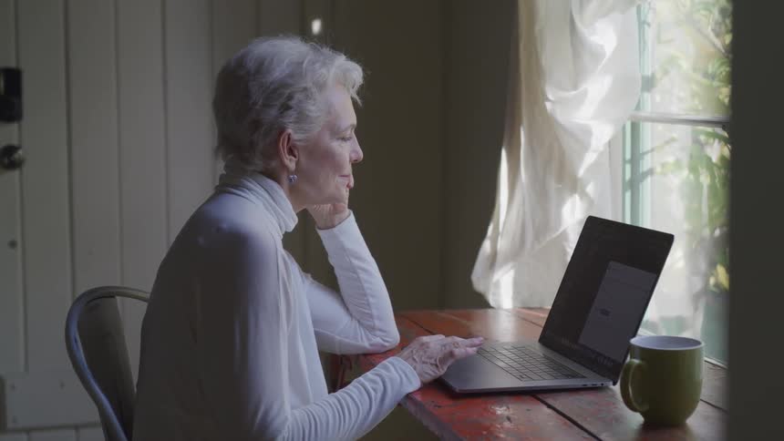 Older woman sits at near window using viewing laptop screen