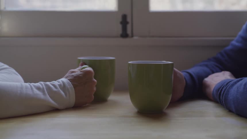 Old man and woman sitting at the table hold green mugs next to the window