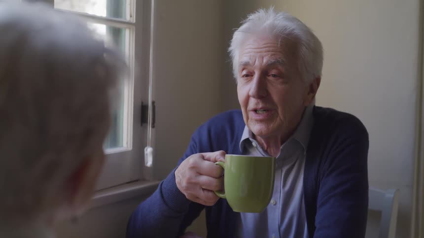 Elderly man holds a mug as he talks to a woman while he sits
