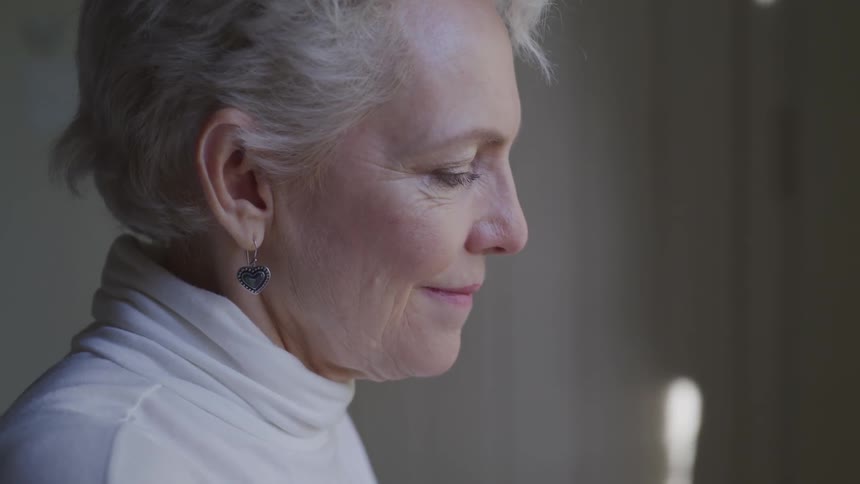 Old woman wearing white pullover looks down and smiles in a room