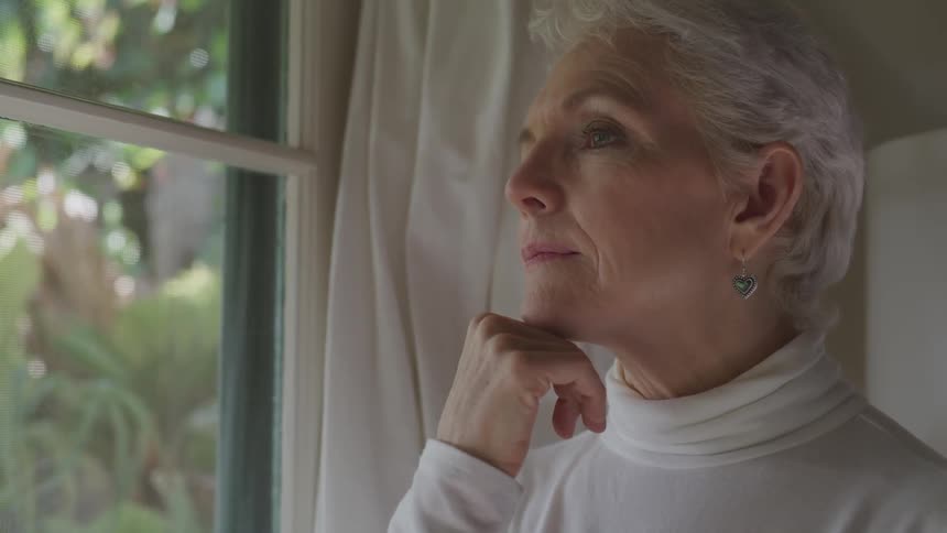 Old woman resting her head on her fingers looks out the window in the bedroom