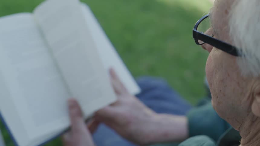 Old woman wearing glasses sits on the grass and reads book in the public park