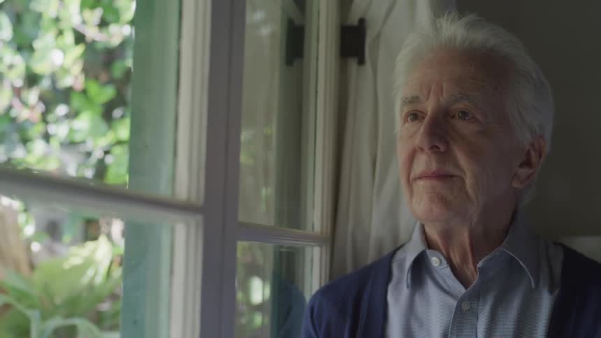 Old man stands and looks out the window in a room during the day