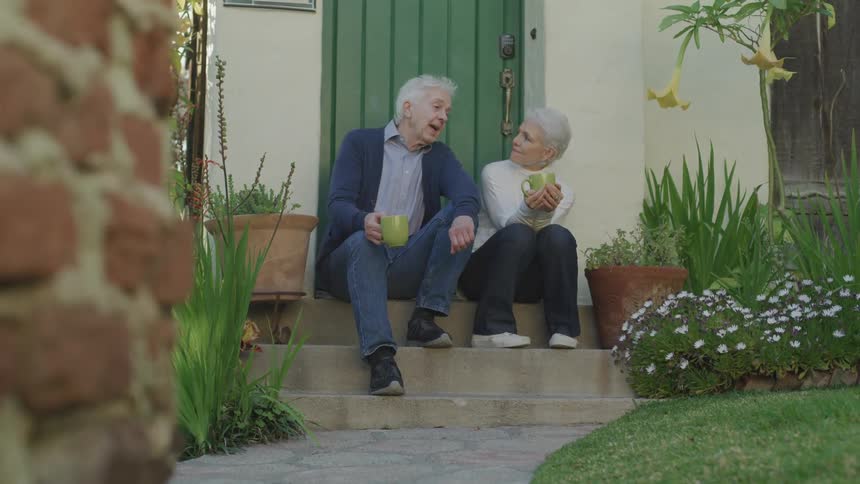 An elderly man and woman sit outside on a step while drinking coffee