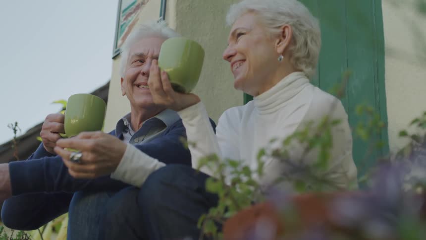 A man and a woman sit on a porch drinking coffee and laughing with each other