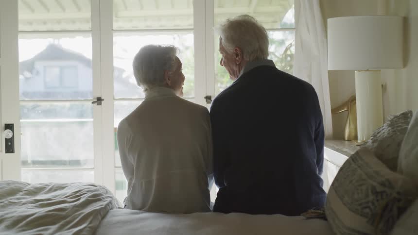An elderly man and woman sit together on a bed and talk