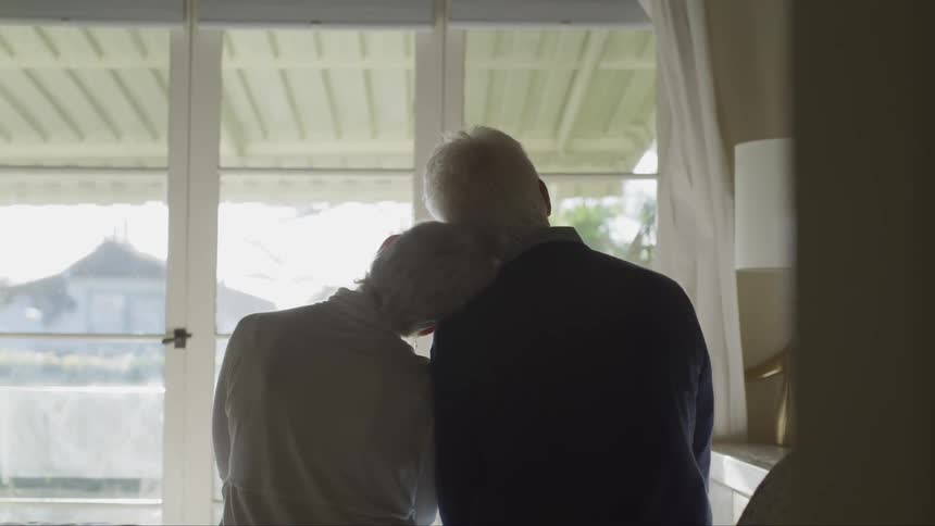 An elderly woman leans her head on a mans shoulder as they look out a window