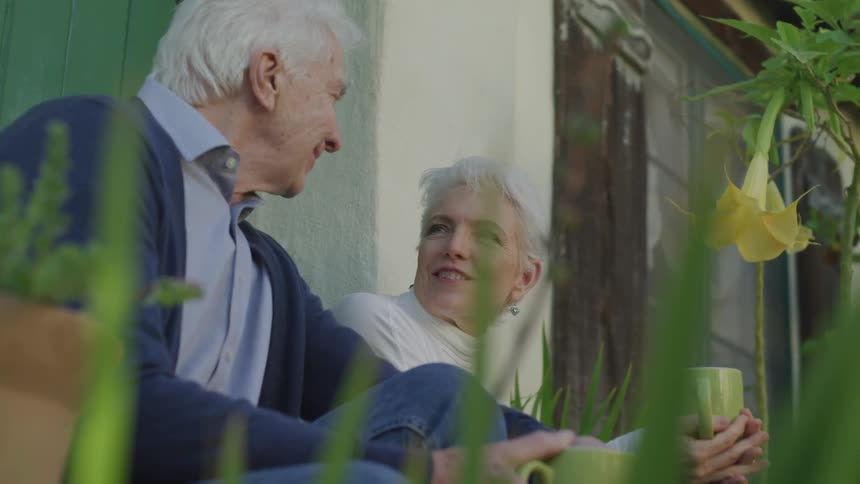 An elderly couple drinks from mugs while chatting on a front step