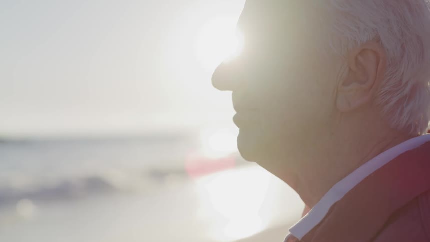 A man with white hair looks contemplative as he looks out over the ocean with the sun shining behind him