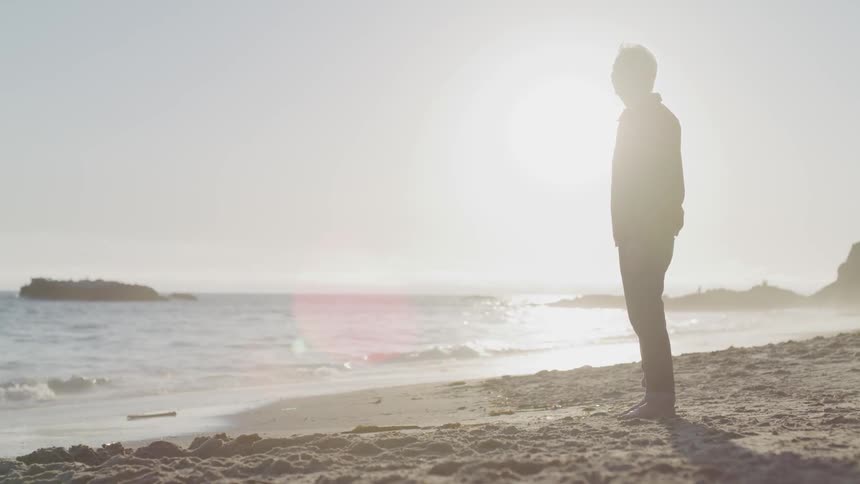 A elderly man stands on the beach and looks out at the water
