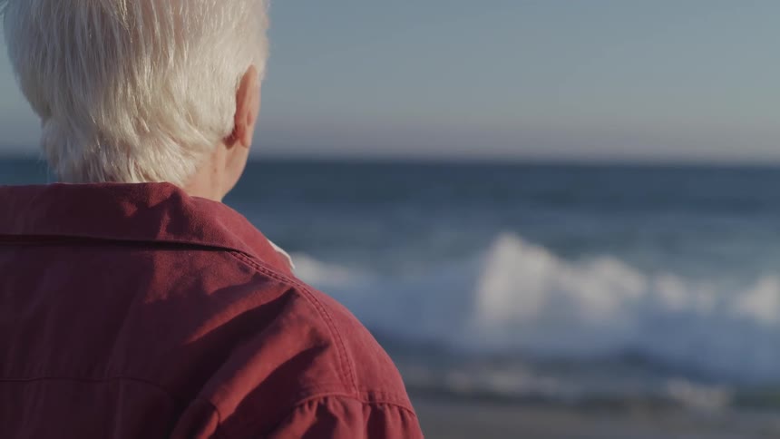 An elderly man stands and looks out at the ocean
