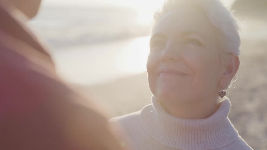 An elderly white woman stands on a beach and gazes up at her spouse