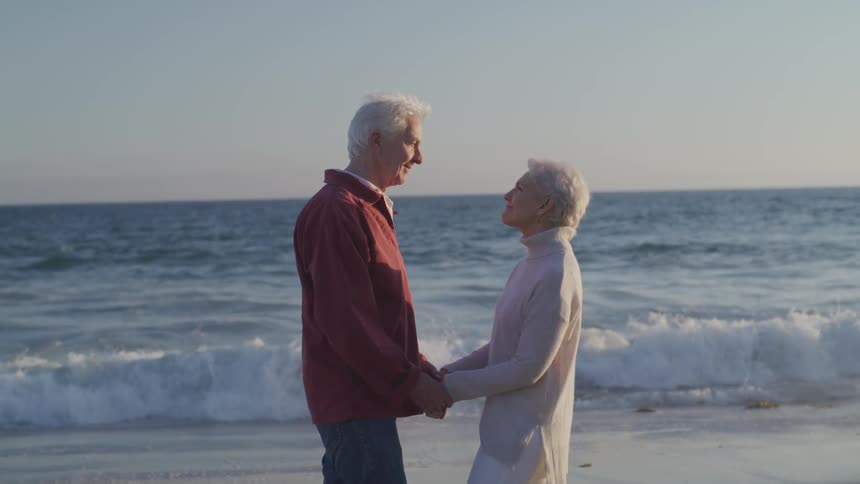 Old man and woman hold hands on the sand beach at sunset next to the ocean