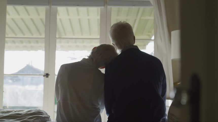 A woman rests her head on her husbands shoulder as they look out a window