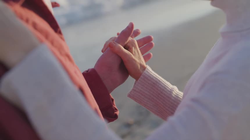 An elderly man and woman hold hands and dance on a beach