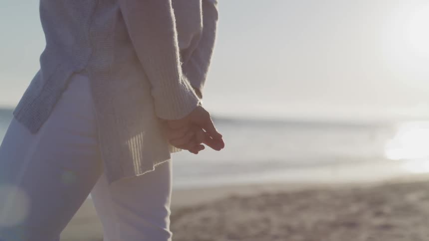 A woman stands on the beach looking out at the water with her hands clasped behind her back