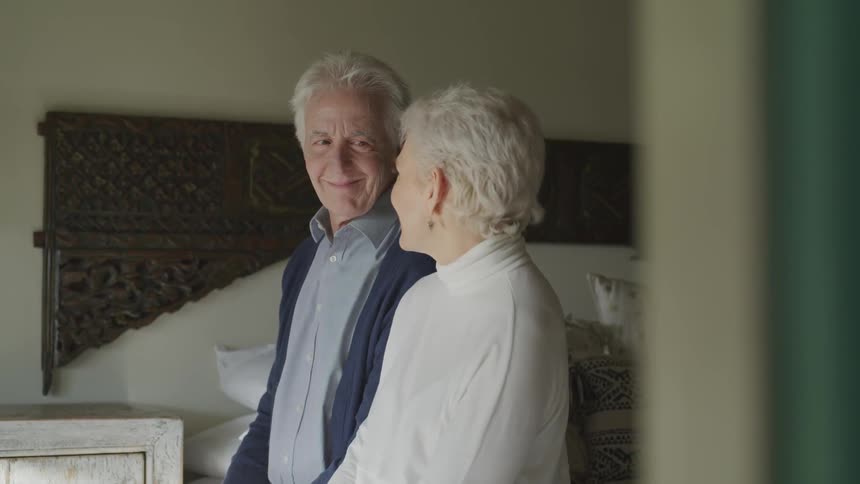 A elderly couple sitting on a bed smile at each other as they have a conversation