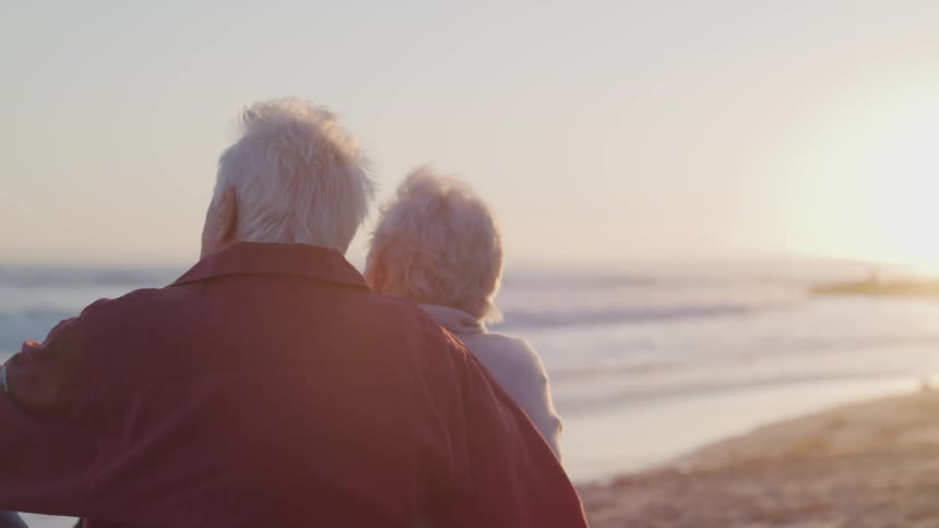 Old man and woman looks at the sea on the sand beach at sunset