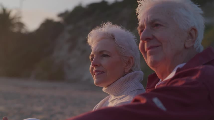 An elderly man and woman sit on the beach together