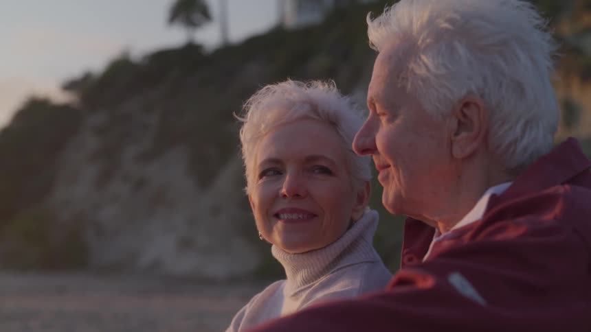 An elderly man and woman sit and look off in to the distance and then smile at one another