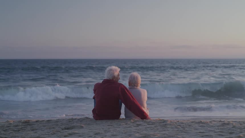 An elderly couple sits together on a beach