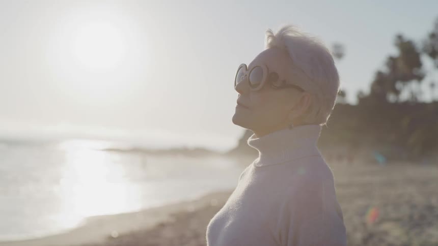 An old woman wearing sunglasses looks out at the ocean as she stands on a beach under the blinding sun
