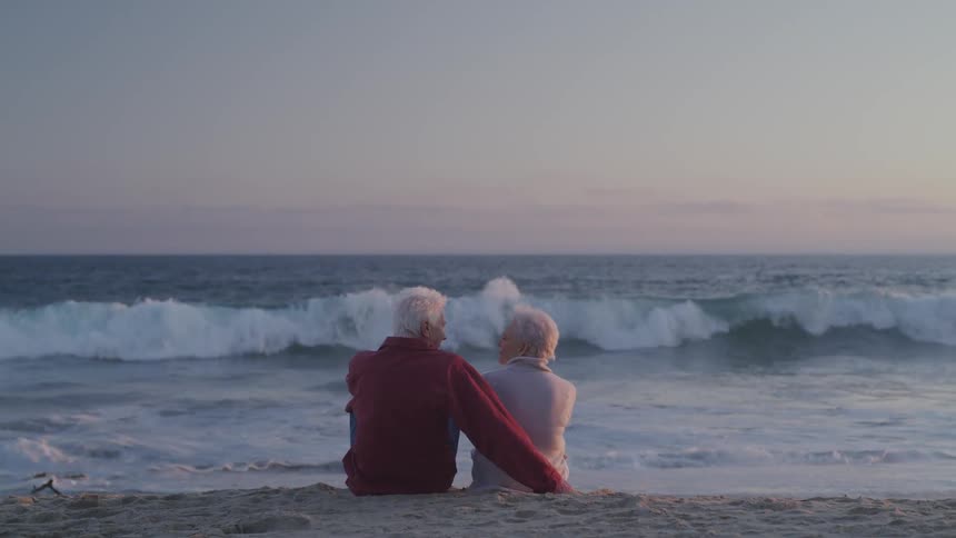 Older couple sits close together on beach at sunset