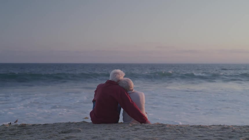 An elderly couple embrace while sitting on a beach