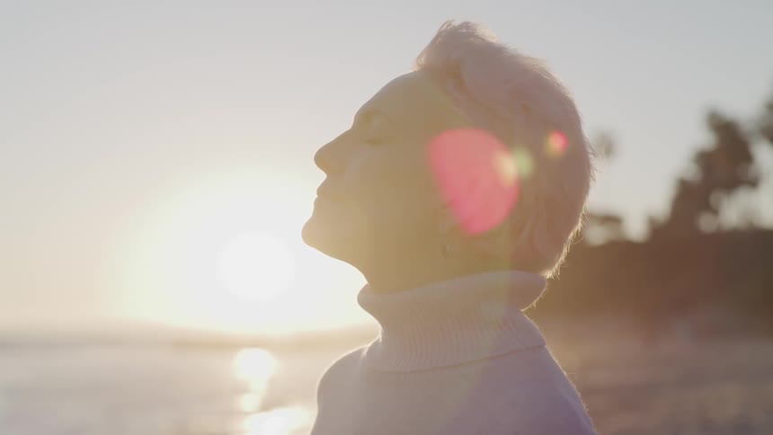 Woman with short gray hair and turtleneck lifts her face up to enjoy a sunrise at the beach