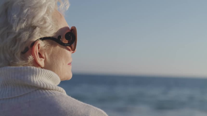 Old woman wearing sunglasses looks at sunseton the beach next to the ocean
