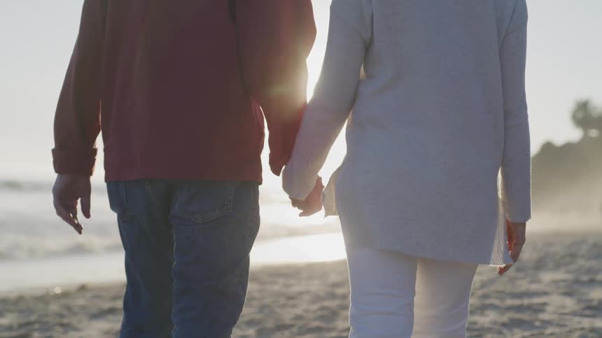A man and woman hold hands as the walk on a sandy beach on a sunny day