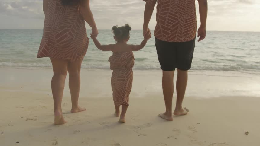 A man and woman walk on the beach while holding the hands of a small child in a dress