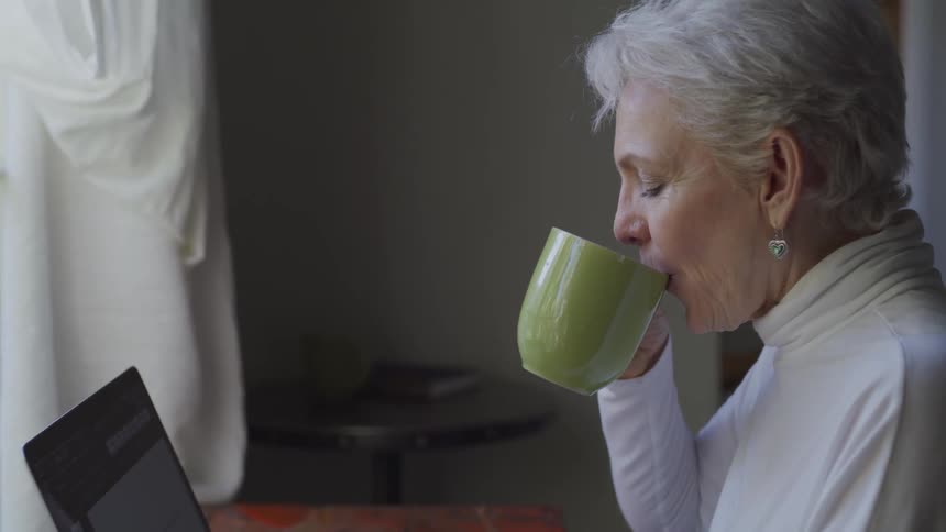 Older woman views laptop near window as she drinks from a mug