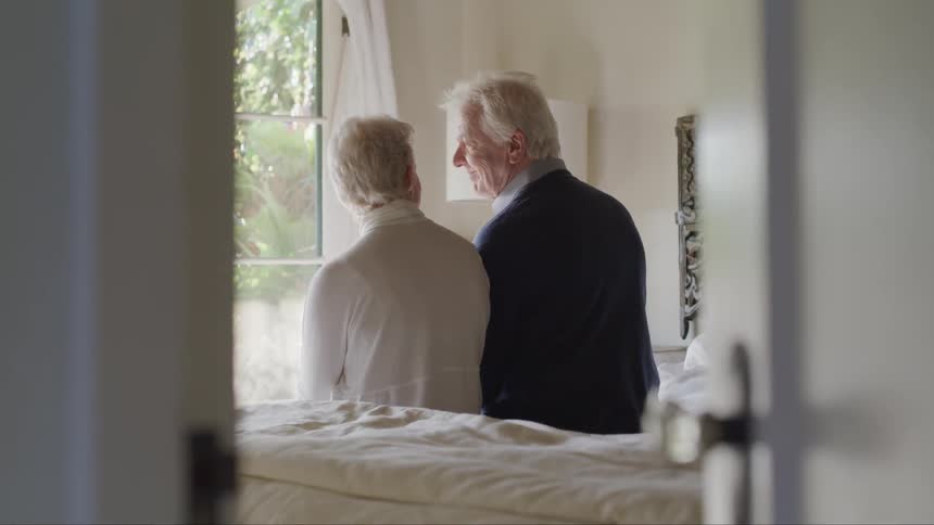 An elderly man and woman sit on a bed together