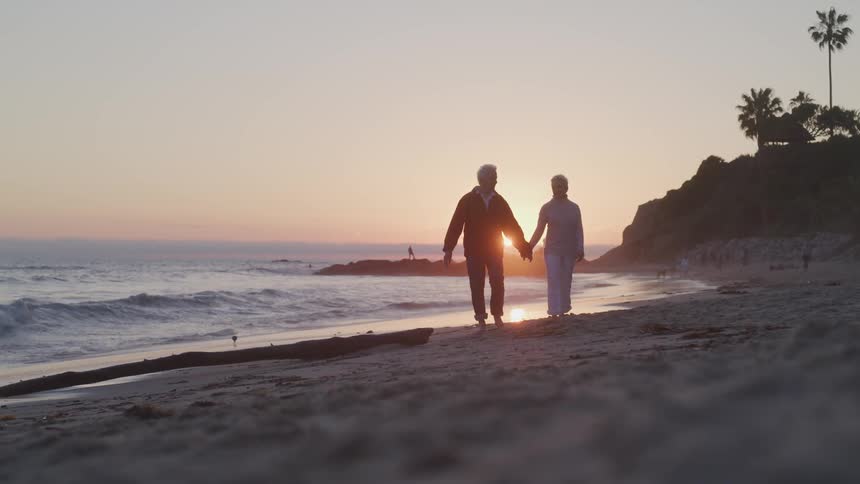 An older couple is holding hands while they walk barefoot down a beach with the sun setting behind them