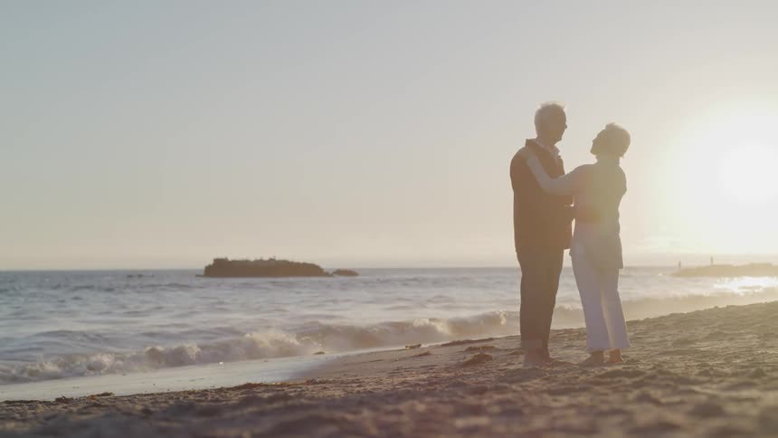 Old man and woman hold each other on the sand beach at sunset next to the ocean