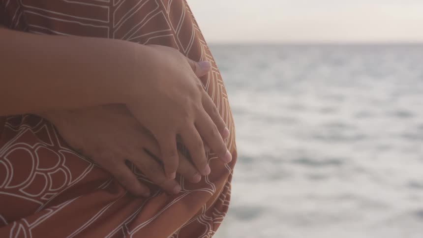 A man and woman stand beside the ocean holding the womans pregnant belly