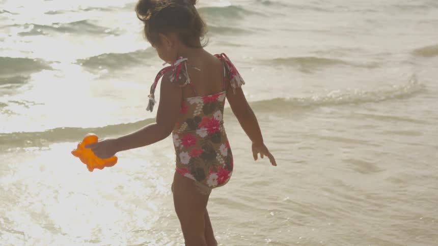 Young female child in a flowered bathing suit runs into the ocean water with a yellow toy in her left hand