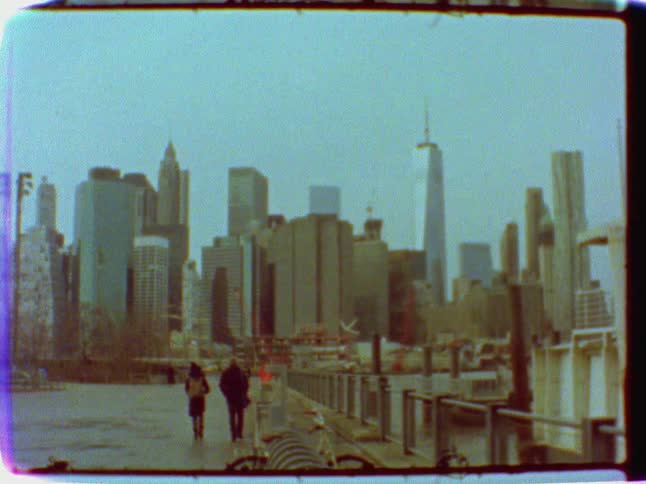 Two people are walking along a river before a large city
