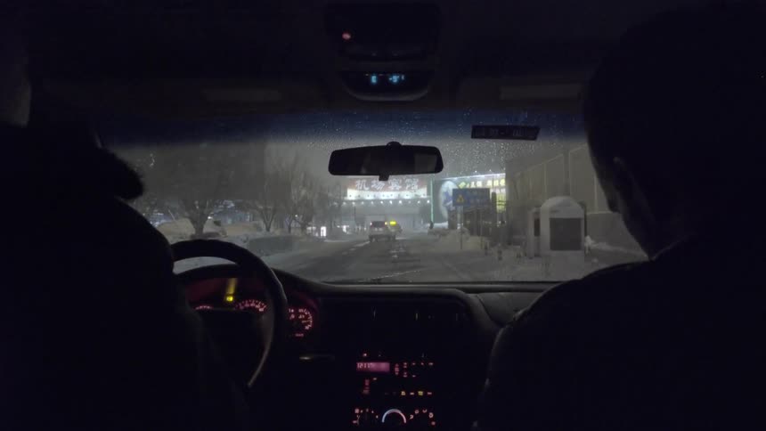 View from inside of a car as a man drives down an urban street at night while it snows