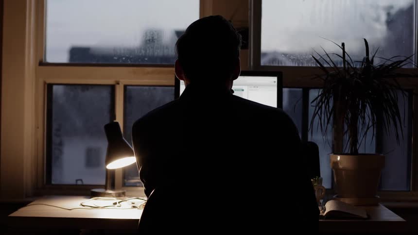 The silhouette of a man can be seen as he sits at a desk near a window in front of a laptop computer
