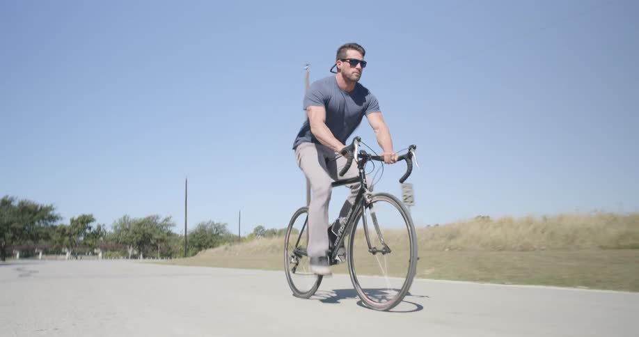 A man wearing sunglasses is riding a bike on a road in a rural area on a sunny day